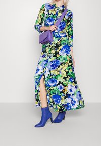 Kukkakuvioinen pitkä mekko vihreillä, sinisillä ja keltaisilla kuvioilla; siinä on kaulus ja sivuhalkio. Yhdistetty violettiin käsilaukkuun ja sinisiin nilkkureihin.