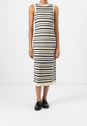 Personne portant une robe crème sans manches, longueur genou, avec des rayures horizontales noires et des mocassins noirs, debout devant un fond blanc uni.