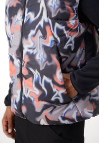 Pufjakas ar abstraktu melnu, pelēku, sarkanu un zilu rakstu. Aprīkots ar priekšējo rāvējslēdzēju un sānu kabatām, izgatavots no viegla, spīdīga materiāla.