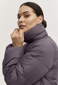 Puffer bunda v tlumené levandulové barvě, s vysokým límcem a prošívaným designem. Modelka jemně táhne límec, aby ukázala texturu a barvu.