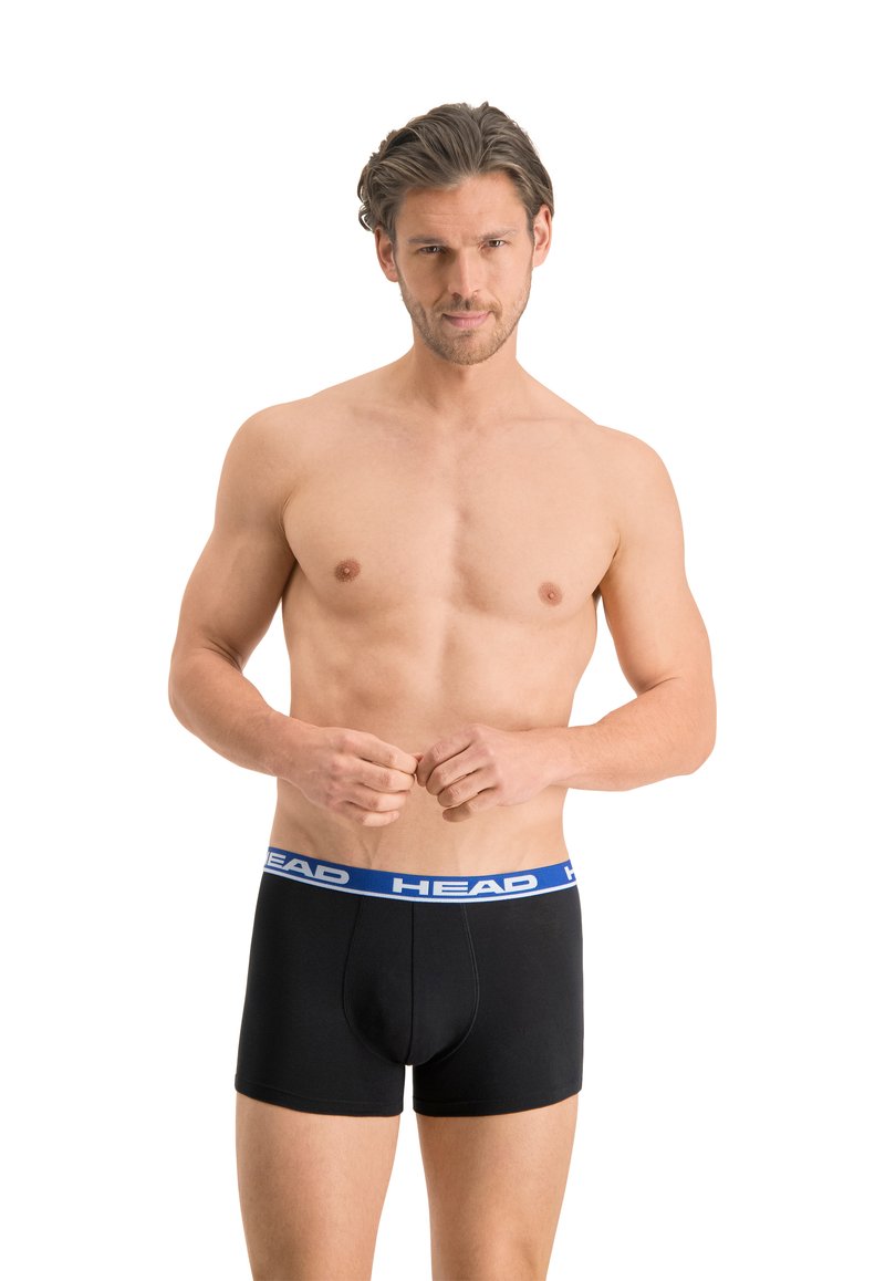 Boxers noirs avec une ceinture élastique bleue portant la marque "HEAD", offrant un ajustement serré et une texture lisse. Le modèle se tient torse nu, les mains jointes.