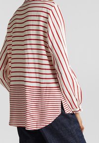 Chemise à rayures rouges et blanches avec des manches longues et amples, des fentes latérales et un ourlet incurvé. Confectionnée en tissu léger avec une coupe décontractée.