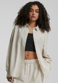 Lys beige oversized skjorte med krage, laget av et mykt stoff. Brukt over en svart crop top, med lange ermer og en avslappet passform.
