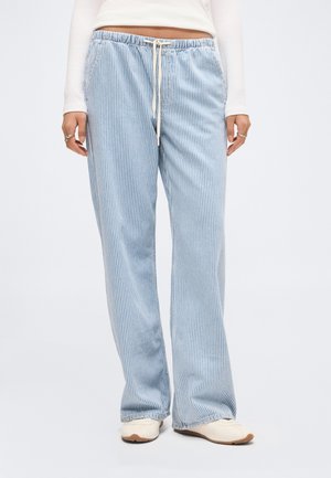 Osoba nosząca luźne, jasno-niebieskie i białe spodnie w pionowe paski z wiązanym pasem oraz białe trampki.