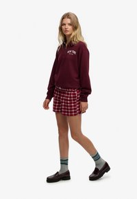 Bordowa bluza z suwakiem do połowy z napisem „New York”, zestawiona z czerwonymi szortami w kratę. Dodatki to szare skarpetki w paski i ciemne buty.
