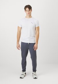 Hvid kortærmet sportshemd med et gråt logo, kombineret med navyblå joggers med grøn detalje ved taljen og hvide sneakers.