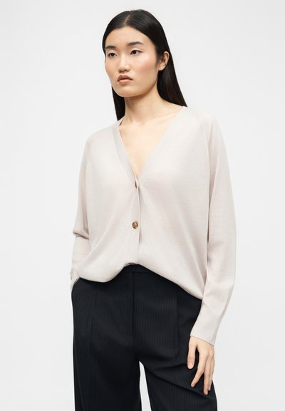 Νεαρή γυναίκα με ίσια σκουρόχρωμα μαλλιά, ντυμένη με χαλαρό μπεζ ζακέτα με κουμπιά και μαύρο παντελόνι, μπροστά σε απλό φωτεινό φόντο.