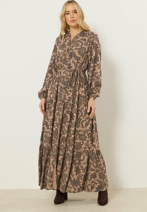 Donna con capelli lunghi biondi che indossa un maxi abito beige a fiori con bottoni e stivali marrone scuro alla caviglia, in piedi contro un muro semplice.