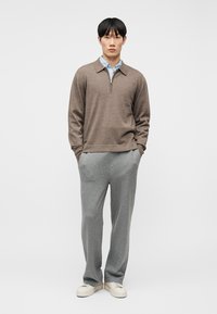 Jeune homme debout les mains dans les poches, portant un pull marron à col zippé, une chemise bleue à col, un pantalon gris et des baskets blanches sur un fond blanc.