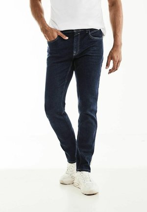 Homme portant un jean slim bleu foncé et des baskets blanches, une main dans la poche et une chemise blanche partiellement visible.