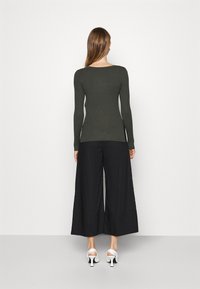 Eine Frau, die von hinten zu sehen ist, trägt ein eng anliegendes, dunkelgrünes, geripptes Langarmoberteil, schwarze, weit geschnittene, knöchellange Hosen und weiße, Absatzsandalen.
