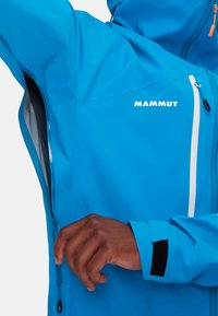 Blaue wasserdichte Jacke mit seitlichem Reißverschlussfach, verstellbaren Manschetten und kontrastierendem weißen Markenlogo. Glatte Textur und modernes Design.