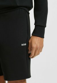 Mustat shortsit, joita on valmistettu sileästä kankaasta. Niissä on herkkä "BOSS"-logo valkoisena vasemmalla puolella, elastinen vyötärö ja rento istuvuus.