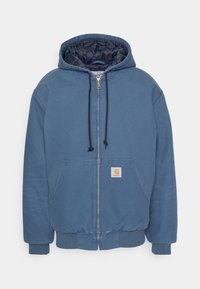 Blauwe hoodie met rits, gemaakt van zachte stof. Heeft een gevoerde binnenzijde, vo pockets, en een klein oranje logo op de linker pocket.