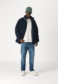 Marinblå fleece-jakke, hvit t-skjorte, lyseblå jeans og hvite sneakers. Grønn cap med logo. Har en kjedehalskjede og en avslappet passform.