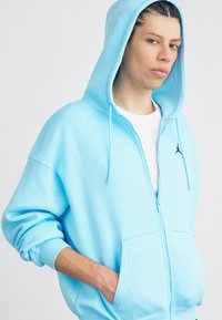 Ανοιχτό μπλε ζακέτα με φερμουάρ από μαλακό ύφασμα, με μπροστινή τσέπη και κορδόνια, με μικρό μαύρο λογότυπο Jumpman στο στήθος.