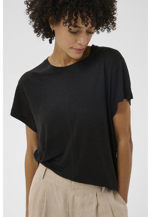 Kobieta o kręconych włosach ubrana w luźną czarną bluzkę z krótkim rękawem, wpuszczoną do beżowych spodni z wysokim stanem, patrząca w dół.