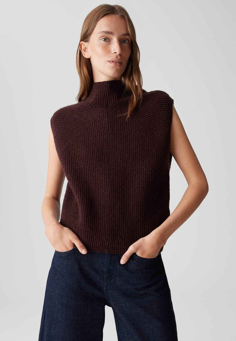 Top senza maniche a collo alto in un intenso bordeaux con texture a coste, caratterizzato da una vestibilità ampia e un design delle spalle angolare. Abbinato a pantaloni in denim scuro.