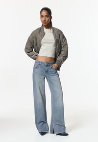 Siva bomber jakna, svetlo siva kratak majica s utisnutim tekstom, široke plave farmerke s blagim izbleđenjem, elegantna crna obuća.