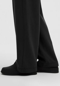 Schwarze Sportschuhe mit abgerundetem Zehenbereich, glatter Textur und minimalistischem Design, kombiniert mit lockeren schwarzen Hosen.