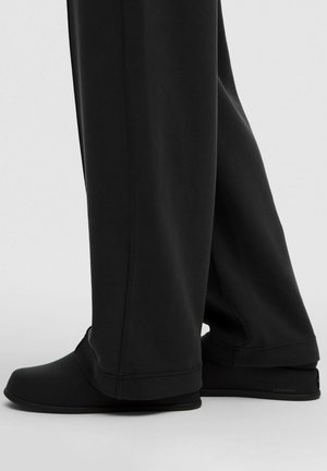 Chaussures de sport noires avec un bout arrondi, une surface lisse et texturée, et un design minimal, associées à un pantalon noir ample.