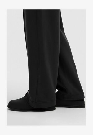 Chaussures de sport noires avec un bout arrondi, une surface lisse et texturée, et un design minimal, associées à un pantalon noir ample.