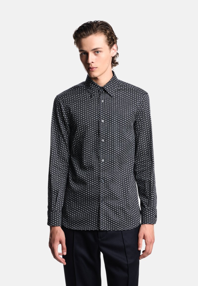 Camicia nera a maniche lunghe con un motivo geometrico bianco, chiusura a bottoni, colletto e tessuto texturizzato. Abbinata a pantaloni scuri.