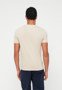 Μπεζ κοντομάνικο μπλουζάκι με στρογγυλή λαιμόκοψη και λεία υφή, που δείχνει τη πίσω όψη και έχει ελάχιστες ραφές, συνδυασμένο με σκούρες βερμούδες.