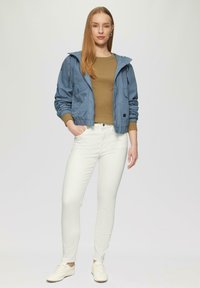Lichtblauwe jas met capuchon over een bruine top, gecombineerd met witte skinny jeans en witte sneakers. Bevat elastische manchetten en zakken.