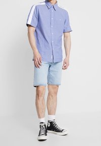 Světle modrá košile s krátkým rukávem a knoflíky, s bílými akcenty na ramenech, spárovaná se světlými džíny a černými teniskami se zvýšeným podpatkem.