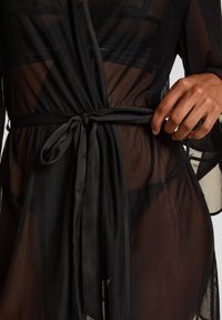 Διάφανο μαύρο ρόμπα δεμένη στη μέση με φαρδιά σατέν κορδέλα, που αποκαλύπτει μαύρα εσώρουχα, το χέρι ακουμπά κοντά στον δεσμό στη δεξιά πλευρά.