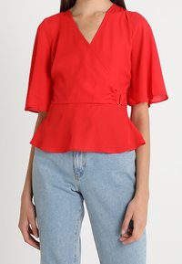 Kvinde iført en rød wrap-bluse med flagrende korte ærmer og peplum-kant, parret med lyseblå højhalset jeans.