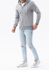 Hoodie gris à fermeture éclair avec une poche kangourou, associé à un jean skinny bleu clair déchiré et des baskets blanches montantes avec des accents gris.