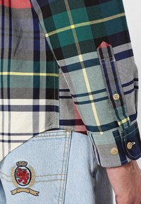 Camicia a scacchi multicolore con tonalità verdi, blu navy e rosse; presenta un colletto a punta e polsini con bottoni; indossata con jeans azzurri chiari con un ricamo sul crest.