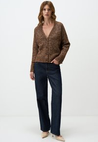 Cardigan en tricot marron avec un décolleté en V profond, six boutons et des poignets côtelés, associé à un jean bleu foncé à jambes larges et des talons beiges.