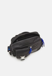 Černá nylonová taška s dvojitými zipy, modrými akcentovými šňůrkami a přední kapsou. Obsahuje stříbrný klip a hladký povrch. Kompaktní, obdélníkový tvar.