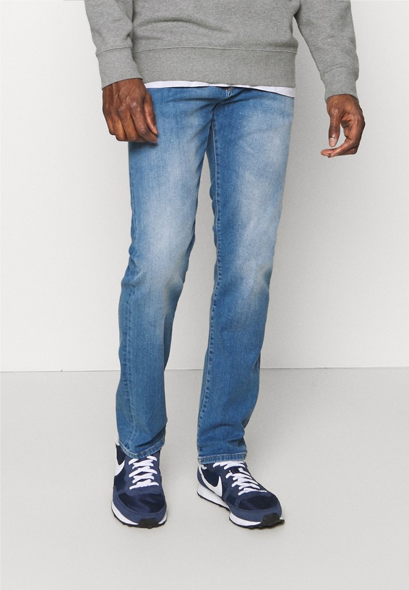 Mies pukeutuneena vaaleansinisiin farkkuihin, tummansinivalkoisiin lenkkareihin ja harmaaseen collegepaitaan, seisoo yksinkertaisen valkoisen taustan edessä.