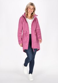 Rozā kapuci ar saites, teksturētu audumu, garām piedurknēm, ar rāvējslēdzēju un divām frontālajām kabatām, apvienota ar tumšiem džinsiem un baltām sporta apaviem.