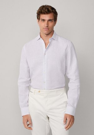 Ein Mann trägt ein helllila gestreiftes Hemd mit Knöpfen, das in weiße Hosen gesteckt ist, und steht vor einem schlichten grauen Hintergrund.