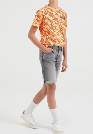 Niño con camiseta de patrón de camuflaje naranja, pantalones cortos de denim gris arremangados, calcetas blancas y zapatillas blancas, de pie con una mano en el bolsillo.
