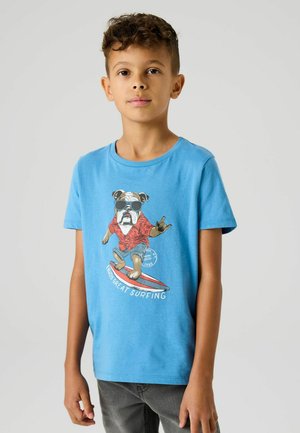 Chlapec v modrom tričku s grafikou buldočka v slnečných okuliaroch, ktorý surfuje na červeno-bielej doske, stojaci pred jednotným pozadím.