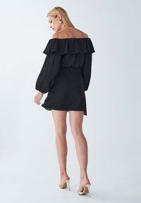 Čierne off-shoulder šaty s volánikovými detailmi, dlhými priesvitnými rukávmi a priliehavým strihom. Nosené s zlatými stiletto vysokými podpätkami.