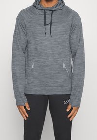 Sudadera con capucha gris, confeccionada en tela texturizada, con bolsillos laterales y cordón. Combinada con pantalones deportivos negros.