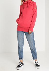 Sudadera roja con capucha, cordones, bolsillo tipo canguro y parche con logo; combinada con jeans de color azul claro y zapatillas negras.