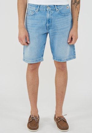 Uomo che indossa pantaloncini di jeans azzurri e scarpe da barca marroni con lacci, in piedi con le braccia rilassate ai lati su uno sfondo bianco semplice.