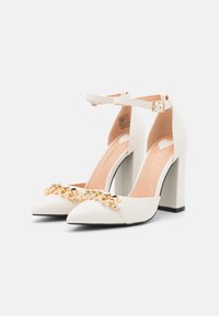 Escarpins blancs à bout pointu avec un détail chaîne dorée sur le devant, bride à la cheville et un talon épais carré. Texture lisse, design minimaliste.
