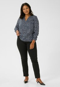 Marinebluse mit cremfarbenem abstraktem Muster, V-Ausschnitt, langen Ärmeln und elastischen Bündchen. Kombiniert mit schwarzen, enganliegenden Hosen und spitzen Slingback-Schuhen.