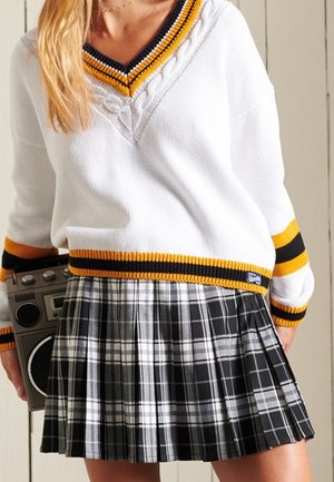 Personne portant un pull blanc à rayures jaunes et noires et une jupe plissée à carreaux noir et blanc tenant un boombox vintage.