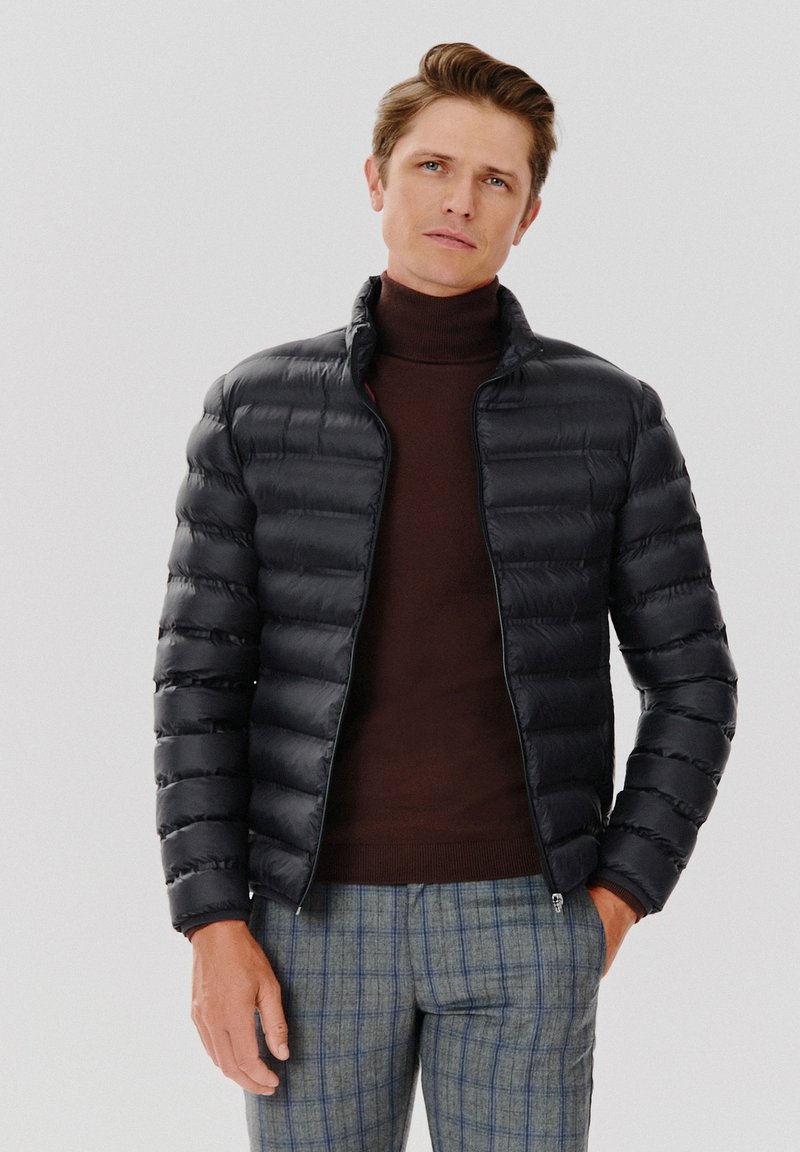 Czarna kurtka puchowa o dopasowanym kroju, pikowana tekstura i wysoki kołnierz, noszona na brązowym golfie, w połączeniu z szarymi spodniami w kratę.