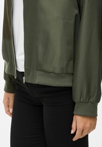 Bomber jacket verde in satin con zip frontale, polsini e fondo a coste, dotato di tasche laterali. Indossato sopra pantaloni neri aderenti.